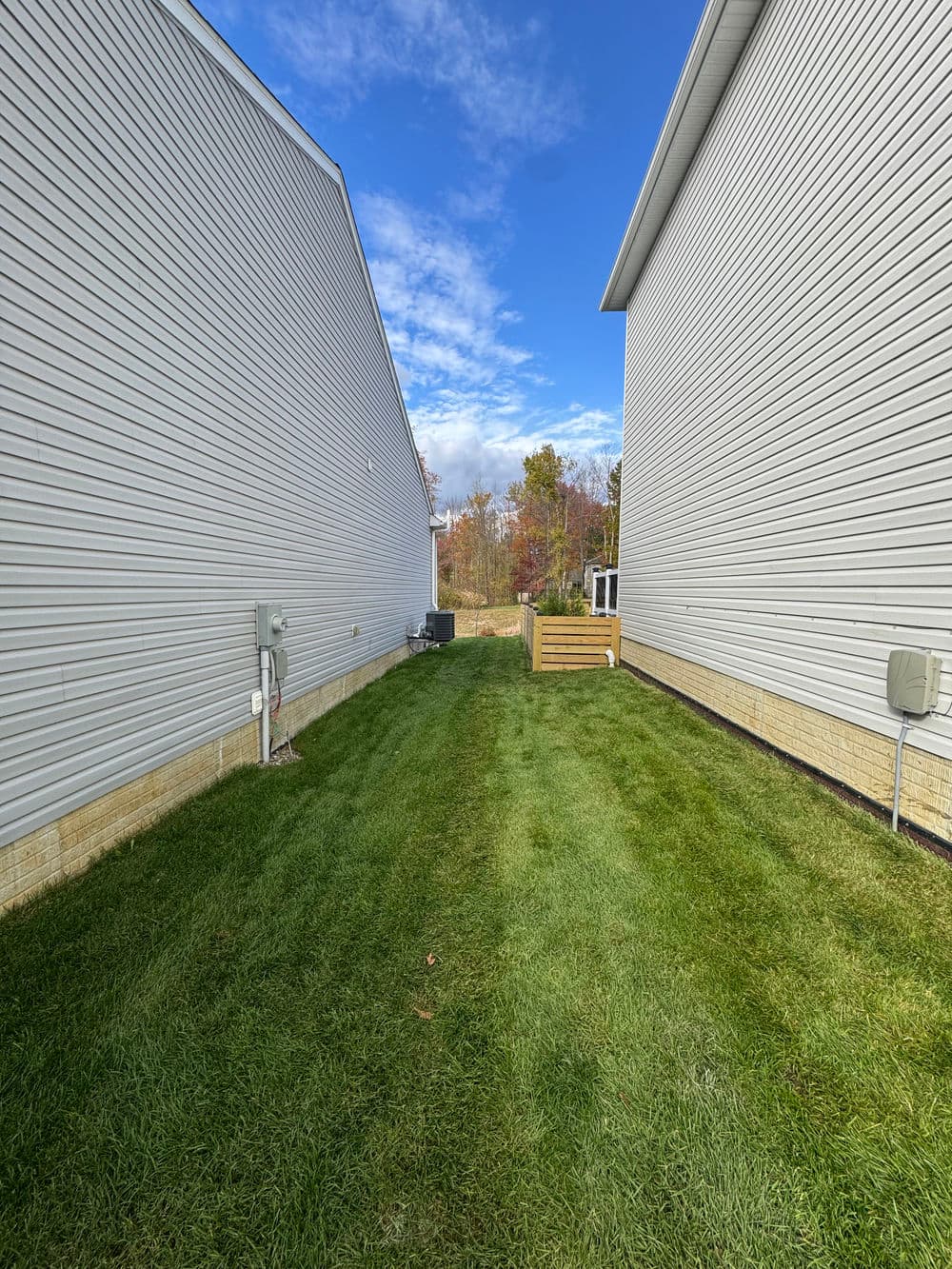 Narrow grassy pathway between two houses with clear sky and trees in the background.
