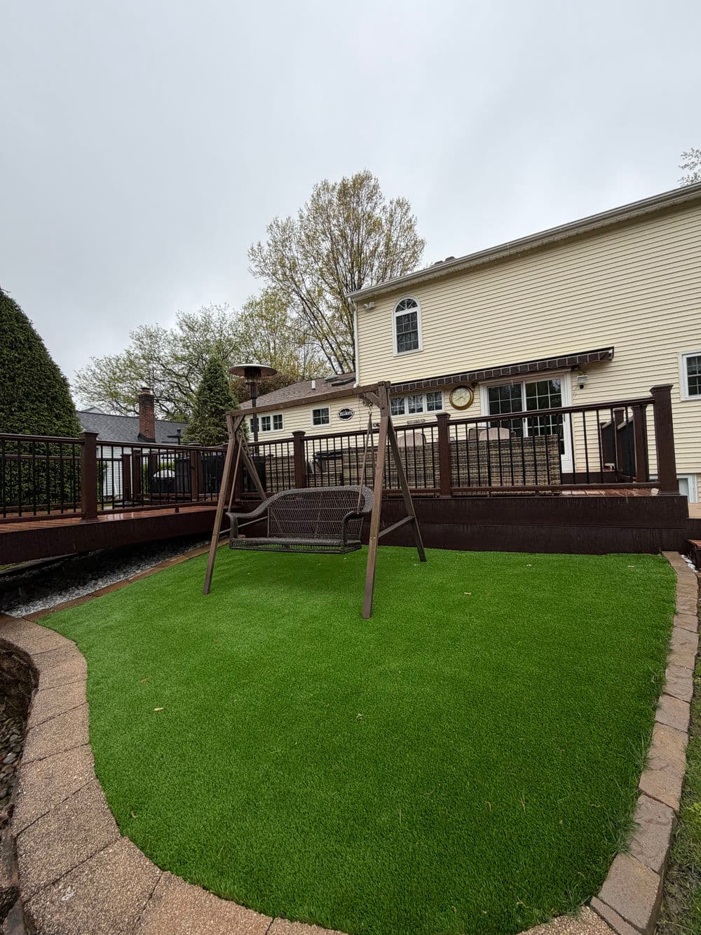 Backyard with artificial grass, swing set, and a view of a two-story house under cloudy sky.
