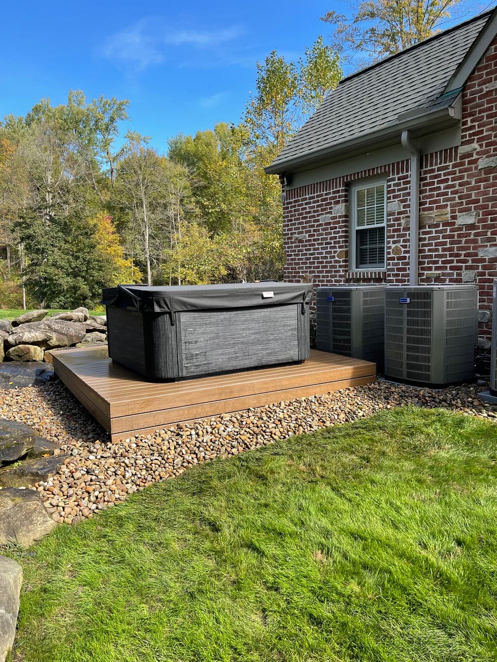 Hot tub on a wooden deck by a brick house, surrounded by gravel and greenery.