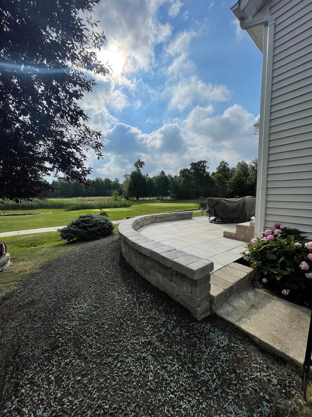 Curved stone patio next to grass and trees under a blue sky and clouds. Garden setting.