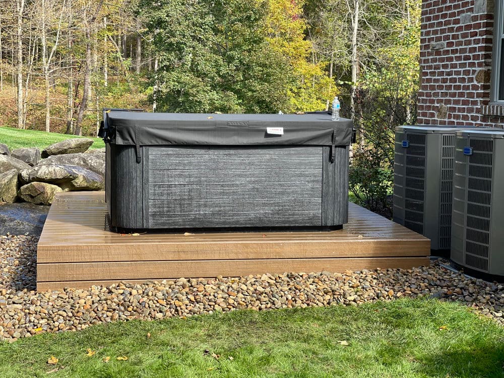 Hot tub on wooden deck surrounded by rocks and greenery, with air conditioning units nearby.