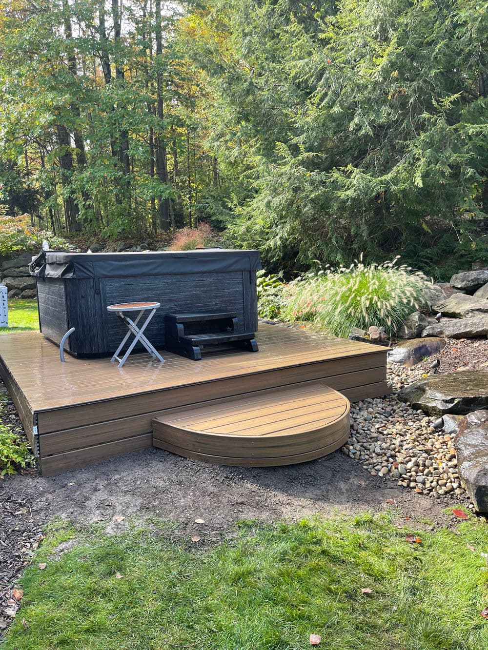 Hot tub on a wooden deck surrounded by greenery and rocks in a tranquil outdoor setting.
