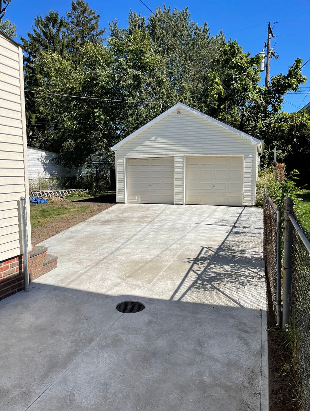 Newly paved driveway leading to a white two-car garage with a fenced yard and greenery.