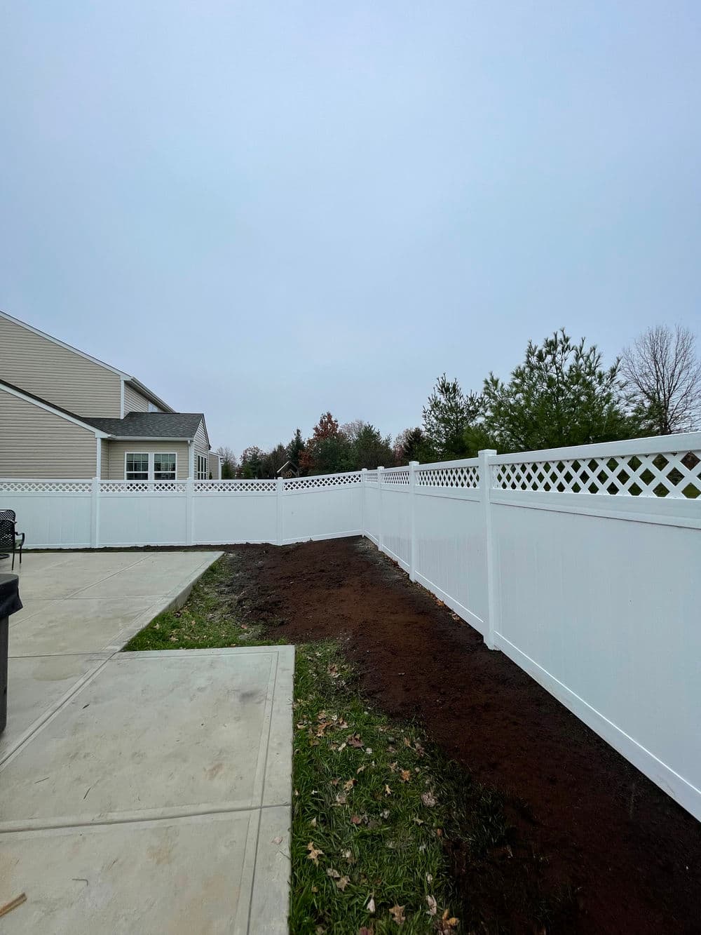 New white vinyl fence along landscaped yard with freshly turned soil and cloudy sky.