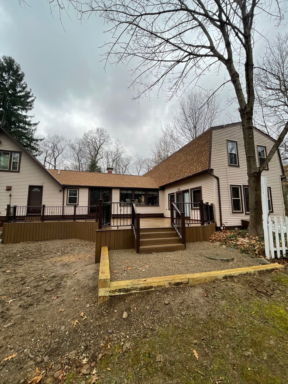 Renovated house with wooden deck, surrounded by trees and a gravel yard on a cloudy day.