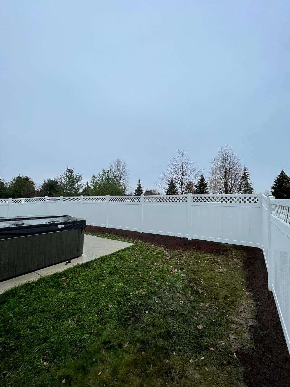White vinyl fence enclosing yard with hot tub under overcast sky and trees in background.