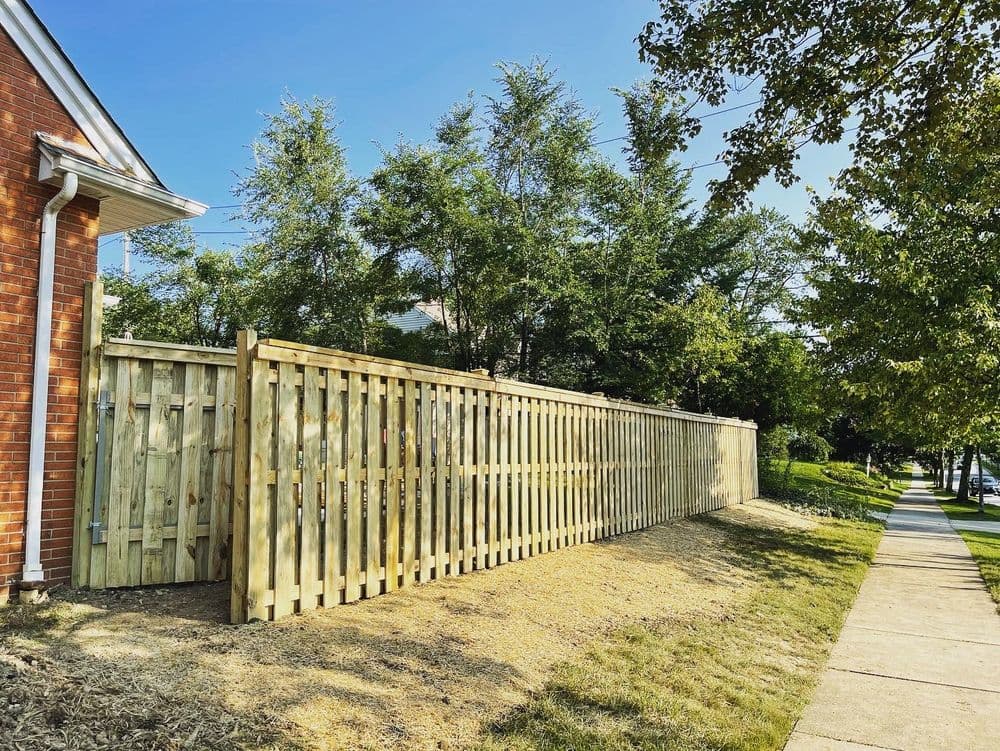 New wooden fence alongside a sidewalk and lush green trees under a clear blue sky.