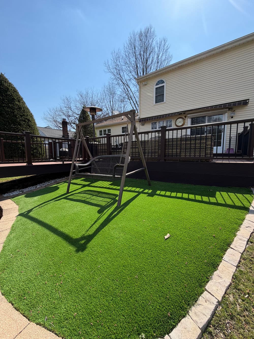 Swing set on artificial grass in a sunny backyard with a deck and clear blue sky.