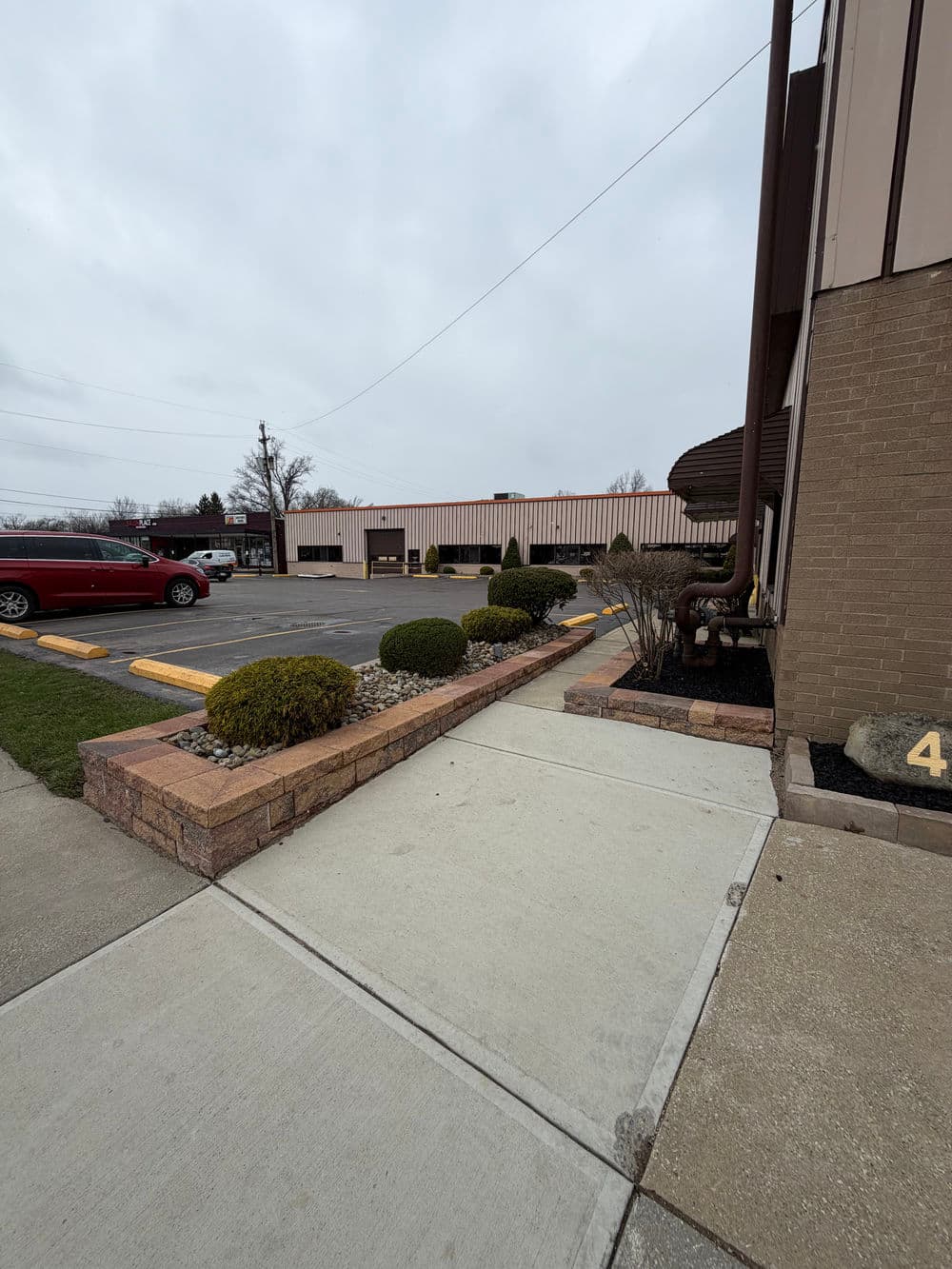 Sidewalk leading to a building with landscaped planters and parking lot on a cloudy day.