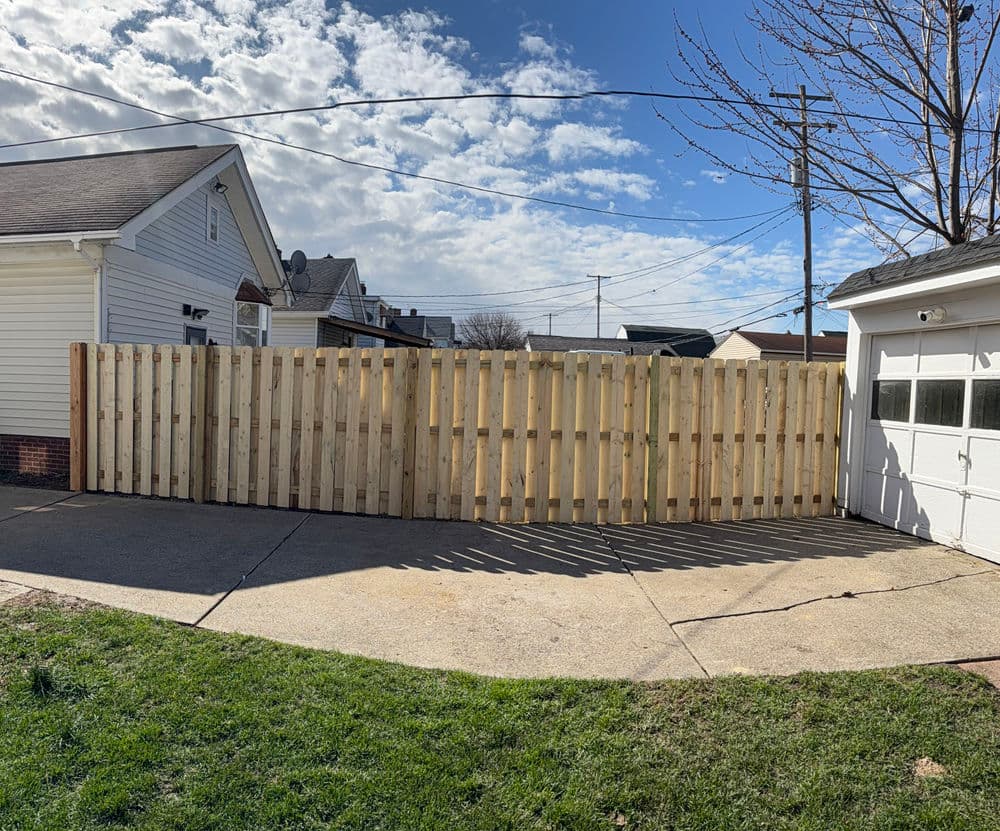 New wooden fence installation in residential driveway under a blue sky with scattered clouds.