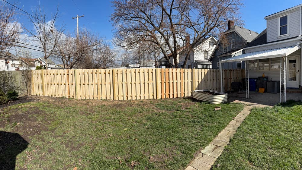Wooden fence enclosing a grassy backyard with a stone path and nearby homes under a blue sky.