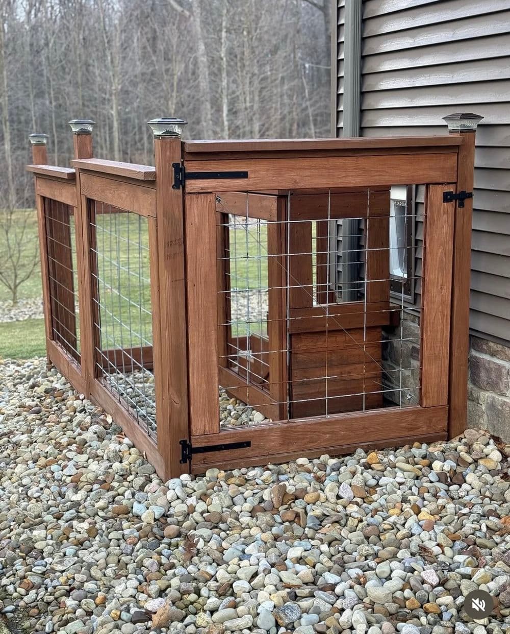 Wooden pet enclosure with wire mesh, located beside a house on gravel landscaping.