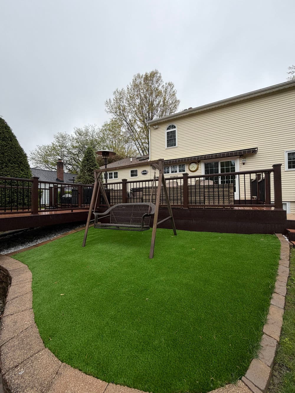 Backyard with synthetic grass, patio, swing set, and two-story house in the background.