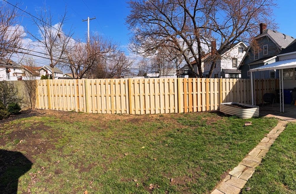 Wooden fence enclosing a backyard with green grass and houses in the background under a blue sky.
