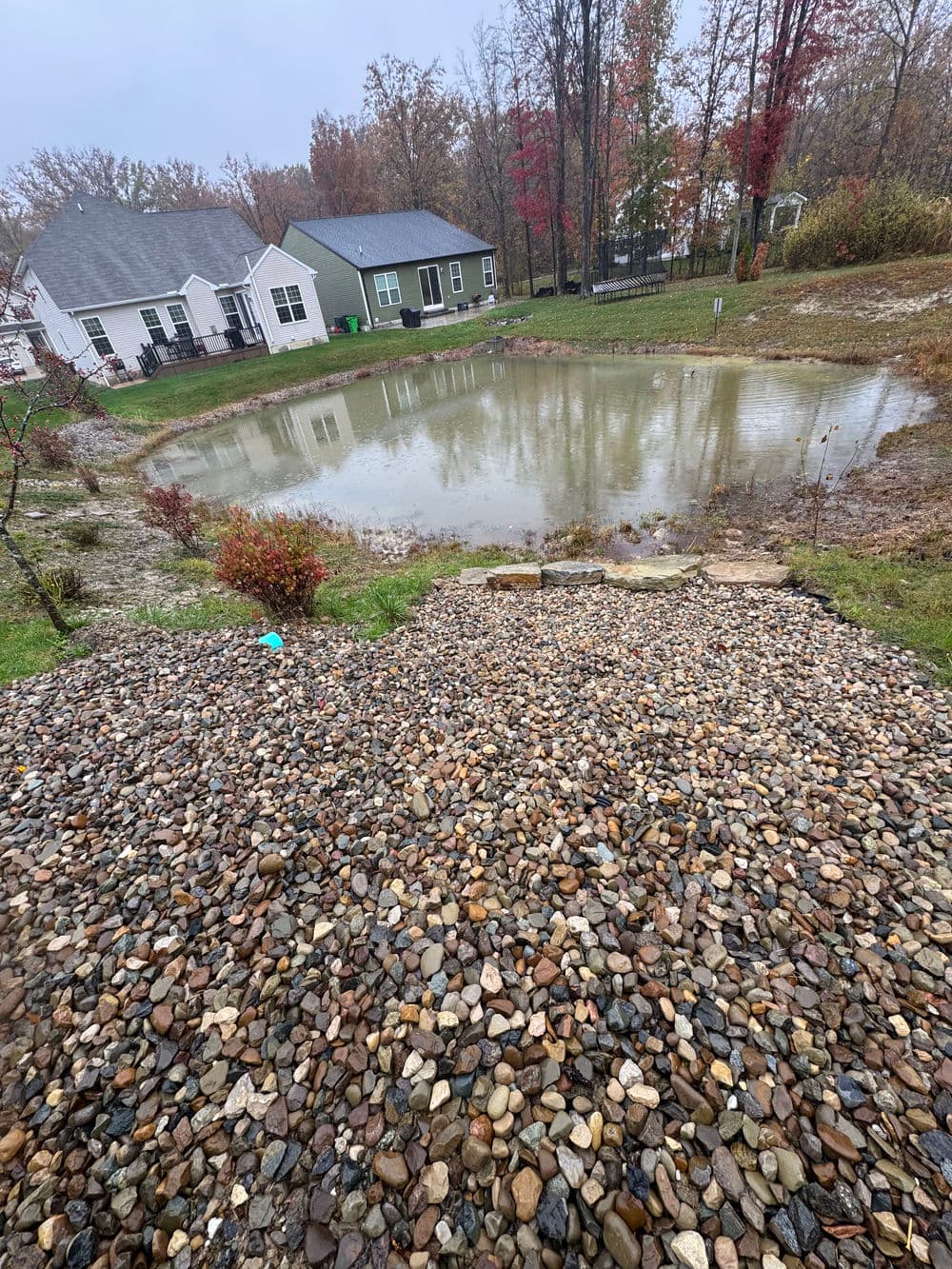 Pond surrounded by rocks and houses, reflecting fall foliage on a rainy day.