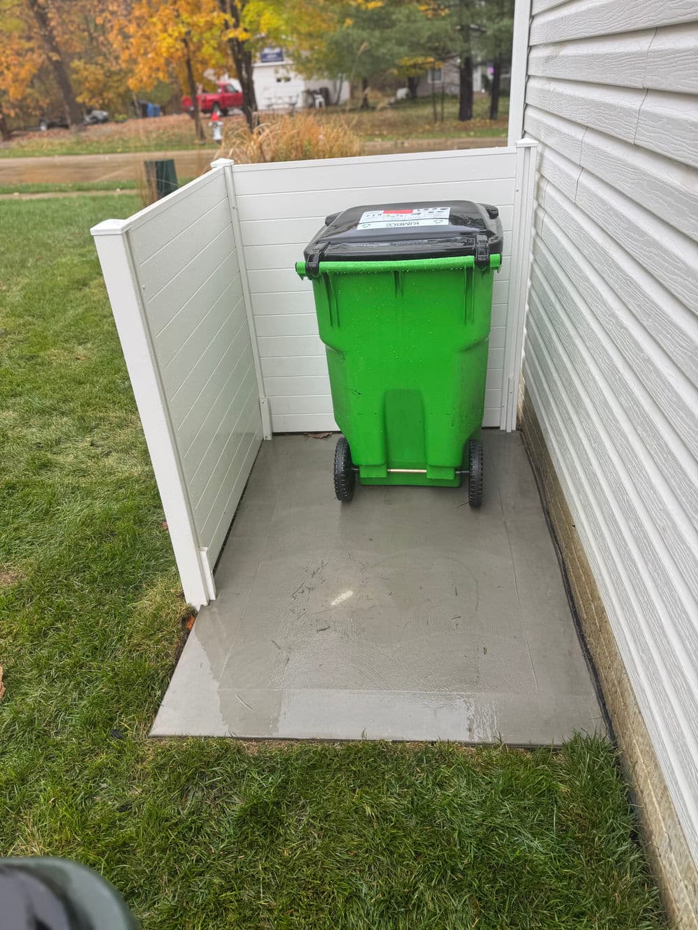 Green trash bin in an enclosed area next to a house on a grassy lawn.